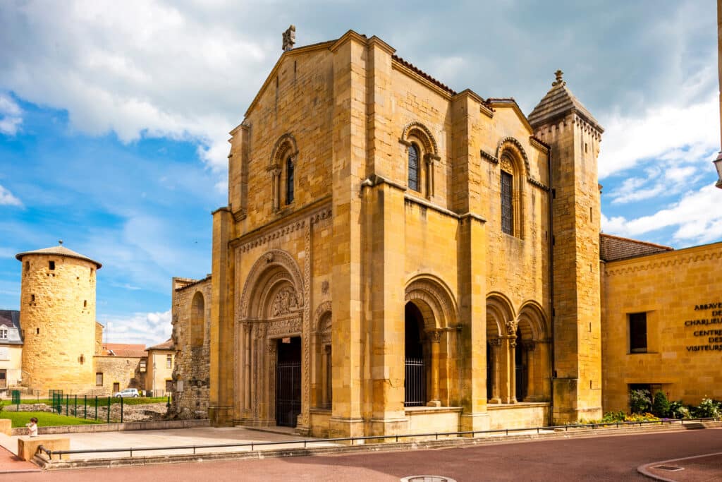 Abbaye romane de Charlieu en pierre jaune, avec une tour ronde et l'entrée sculptée. Panneau 'CENTRE DES VISITEURS'. Ciel nuageux.