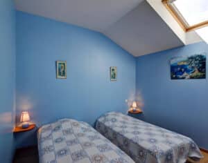 Chambre lumineuse aux murs bleus, deux lits simples avec couvre-lits à motifs. Lucarne au plafond et tableaux décoratifs.