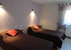 Chambre double lumineuse avec deux lits simples, couvre-lits marron et oreillers orange. Murs lavande, fenêtre et radiateur. Ambiance cozy.