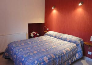 Chambre confortable avec lit double à couette bleue fleurie. Mur rouge texturé, mur beige, bouquet de roses et lumières murales. Radiateur.