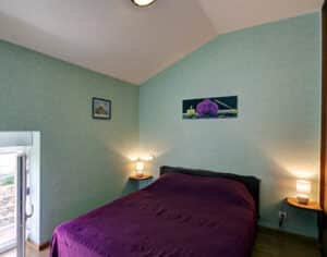 Chambre douillette aux murs verts et lit double violet. Tables de chevet en bois, lampes chaudes et tableaux, dont une orchidée.