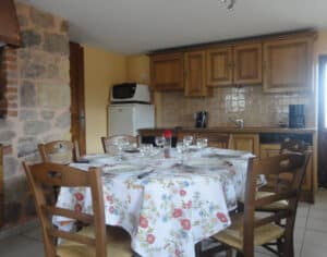 Table ronde dressée avec nappe fleurie et chaises en bois dans une cuisine rustique avec mur en pierre et mobilier bois.
