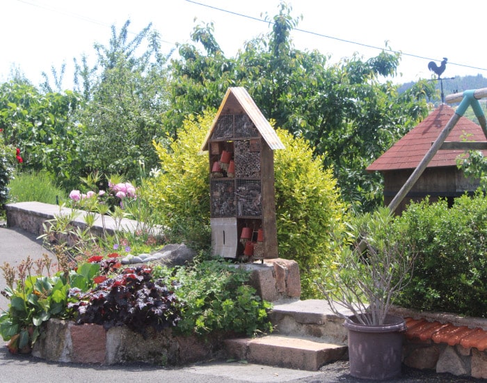 Grand hôtel à insectes en bois dans un jardin verdoyant et fleuri. Des pivoines roses et un coq sur une girouette sont visibles.