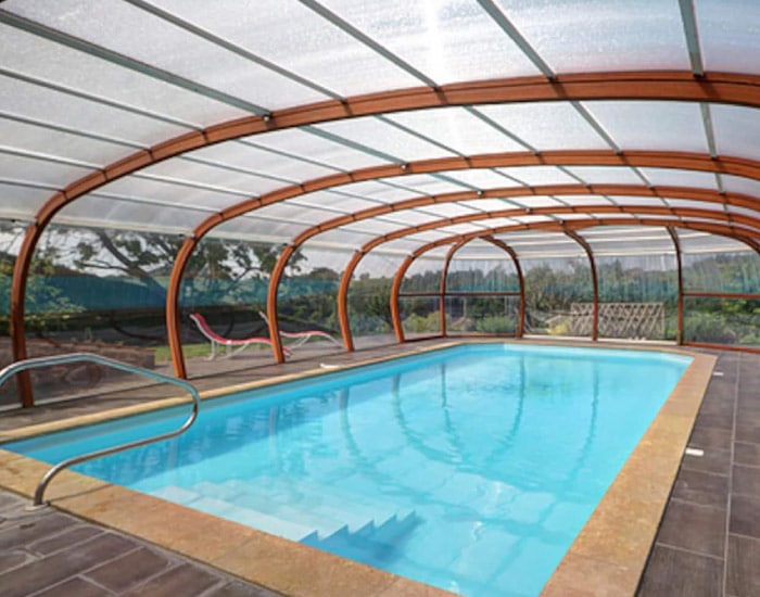 Piscine rectangulaire couverte d'un abri transparent à arceaux bois, avec eau bleue et marches, sur une terrasse carrelée. Vue sur le jardin.